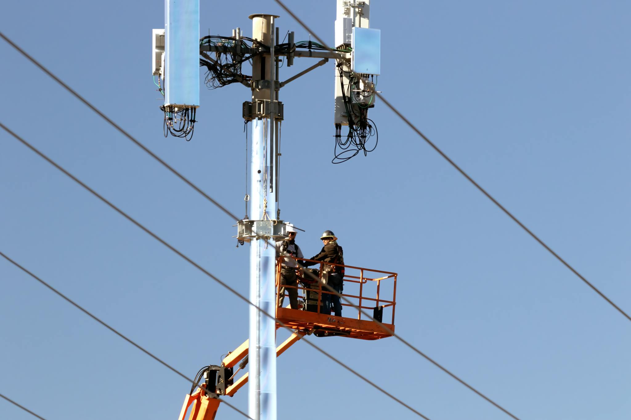 Technicians on a lift installing communication devices on a utility pole against a clear sky.